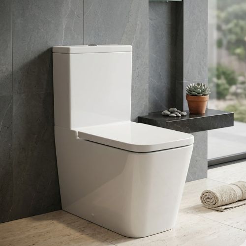 A professional studio shot of the Inspira Square Floor Standing WC, showcasing a white ceramic toilet with a bold, monolithic square design and a soft-close lid, set against a dark grey stone-tiled wall in a minimalist bathroom.