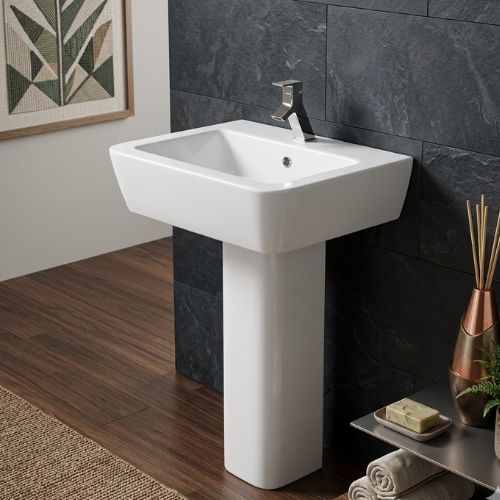 A high-gloss white ceramic Adele semi-pedestal washbasin by Neatflow Solution, wall-mounted against a dark textured stone backdrop with premium spa accessories.