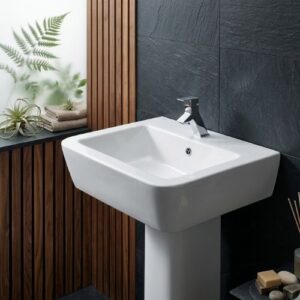 A premium high-gloss white ceramic Advance washbasin by Neatflow Solution featuring a robust full-pedestal design and a modern polished chrome faucet, set against a luxury dark stone and wood-slatted bathroom wall.