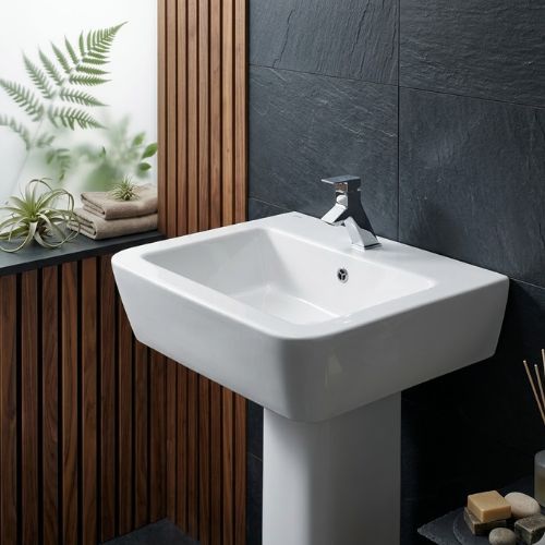 A premium high-gloss white ceramic Advance washbasin by Neatflow Solution featuring a robust full-pedestal design and a modern polished chrome faucet, set against a luxury dark stone and wood-slatted bathroom wall.