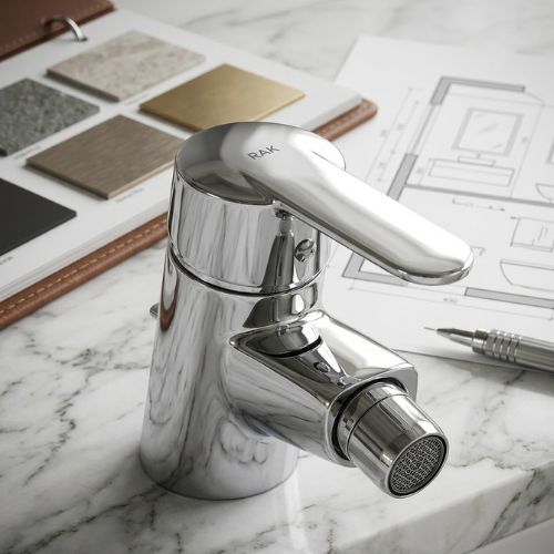 Close-up of a polished chrome bidet mixer tap with an adjustable nozzle, positioned on a white marble vanity next to architectural floor plans and material samples.