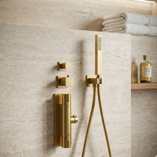 Opulent Aura Gilded Brass thermostatic shower suite in a satin gold finish, mounted on a textured cream stone wall with matching geometric controls and a slim handheld wand.