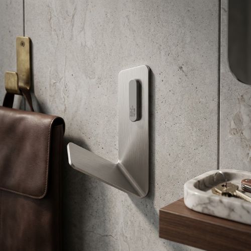 A sleek, L-shaped FLAMA robe hook in brushed aluminium steel, mounted on a grey stone-textured wall alongside high-end leather accessories and marble decor.