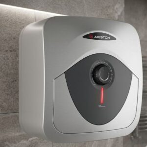 A sleek, wall-mounted Ariston 15L electric water heater with a brushed metallic finish and a glowing red temperature indicator, professionally installed against a modern stone tile bathroom wall.