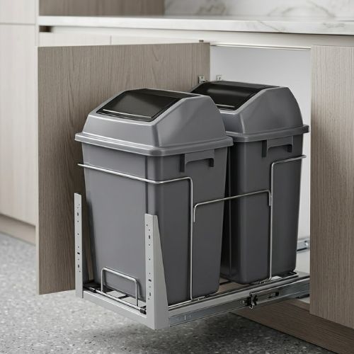 A dual-compartment kitchen pull-out waste bin system installed inside a cabinet, featuring two dark grey bins on metal tracks against a modern wood-paneled kitchen backdrop.