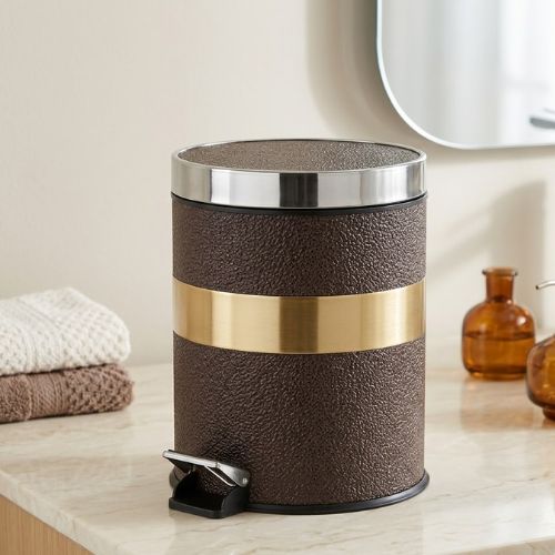 A designer pedal-operated waste bin with a rich brown leather-textured finish and gold-tone metallic accents, placed on a marble countertop in a modern bathroom.