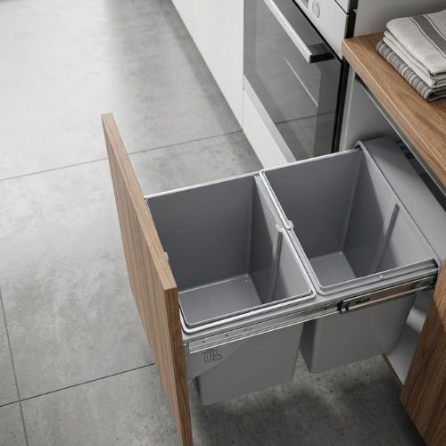 A dual-compartment pull-out waste bin installed inside a wooden kitchen cabinet, featuring a smooth-glide sliding track system.