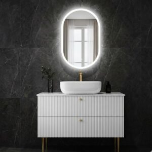 A modern white fluted bathroom vanity with gold-toned legs, topped with a countertop basin and an oval LED-backlit mirror, set against a dark marble wall.