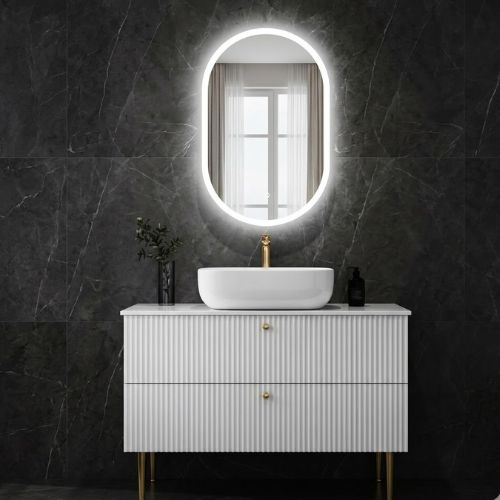 A modern white fluted bathroom vanity with gold-toned legs, topped with a countertop basin and an oval LED-backlit mirror, set against a dark marble wall.