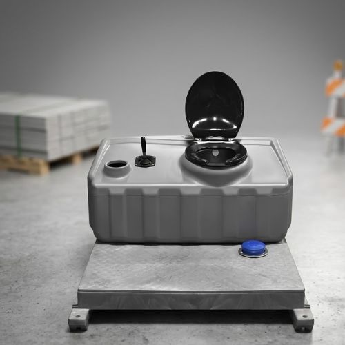 A studio-style shot of a compact, grey mobile flushing toilet unit featuring a black toilet seat and a blue flush button, set against a blurred construction site background with stacked materials