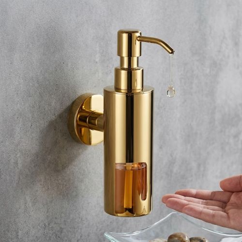 A sleek, gold stainless steel wall-mounted soap dispenser with a visible liquid level, shown dispensing a drop of soap into a hand above a glass bowl of decorative stones.
